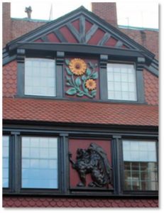 A large wooden carving of a sunflower painted in bright yellow and green hangs under the eave above the front door. Beneath it, in the center of a windowed gallery you see a carving of a black gryphon. 